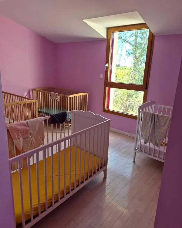 Chambre aux murs roses avec quatre lits à barreaux et une grande fenêtre sur les arbres